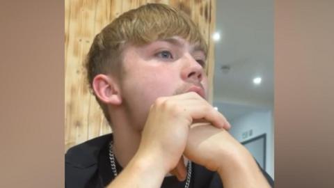A man with blonde hair, in a black top with a silver chain necklace, looking away from the camera.