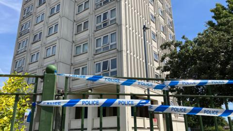Blue and white police tape is tied to a green metal gate. Behind it is a large cream-coloured tower block.