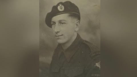 A black and white photograph of a young man in army uniform. He has short dark hair and a beret. 