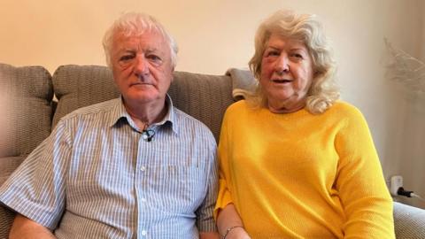 An old man and an old woman sit side by side on a couch. 