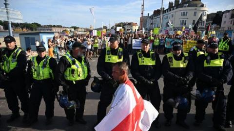 A man draped in a St George flag walks right to left in front of a line of police officers standing behind him and facing forwards, with anti-racism protesters a few metres behind them