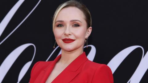Hayden Panettiere wears a red suit and stands in front of a black and white board.