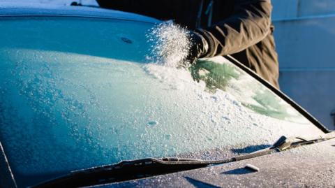 A person, in a black jacket, scraping off frost from a car windscreen