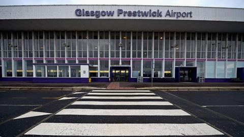 The entrance to a large airport building, with the view coming from across the street. The sign says Glasgow Prestwick Airport