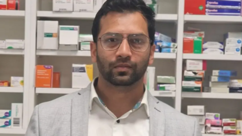 A man with a very short beard is wearing glasses, a white shirt and a grey blazer. He is standing in front of row of medication boxes.