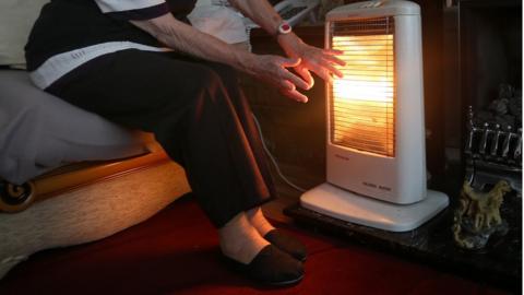 An elderly person staying warm next to a heater