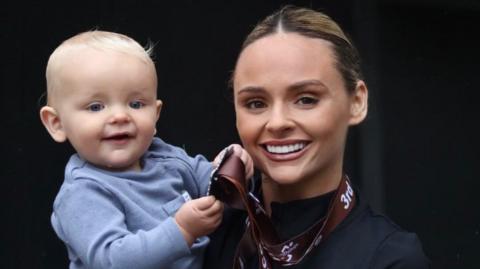 A woman (Chloe) is smiling while holding her blonde haired baby boy. The medal is around her neck, and her baby is holding it in his hands while smiling toothlessly.