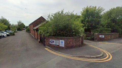 Street view image of the former Xaverian Mission Spirituality Centre in Fulwood. A small red brick building mostly hidden by green trees to the die and back. There are a few cars parked in a car park to the left and the pavements to the side of the building have double yellow lines in a U-shape