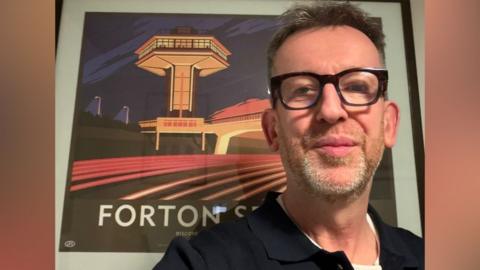 Chris Bradley standing in front of a print of Forton Services. He has short hair, dark-framed glasses and a short greying beard