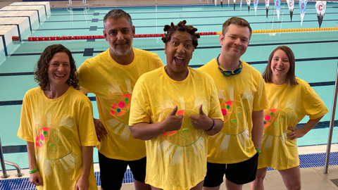 91ȱ CWR presenters and journalists getting ready for their 1,000-mile swimming challenge. The three women and two men are wearing yellow Pudsey bear t-shirts over their swimming costumes, with a swimming pool behind them.