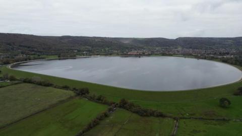 Aerial footage of Cheddar Reservoir, which has green fields in the foreground and the town in the background.