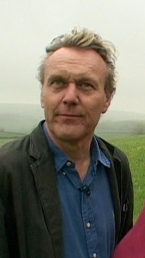 An old picture of Anthony Head pictured outside. A field can be seen behind him. He has short fair hair that's blown back by the wind. He is wearing a blue top and dark jacket.