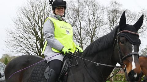Julie-Ann Bowden in a black hard hat, grey sweatshirt and high-vis vest sits smiling on her dark brown hose with a white blaze