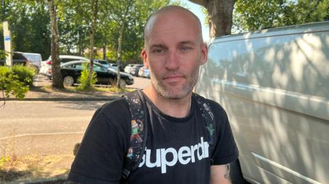 A man is wearing a black t shirt standing under neath a tree.
