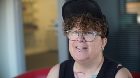 Emma Hastings looks at the camera. She has brown curly hair, wears a cap and glasses and has a lip piercing.