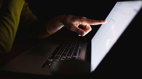 A dark room with a person pointing at a laptop.