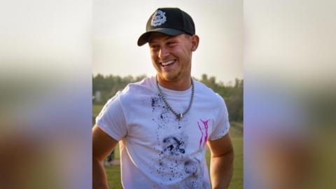 White man wearing a baseball cap and a white t-shirt. He is looking to the right and smiling.