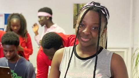 Stephanie Horacio from the Reggae Roots choir looking at the camera, wearing braids and a grey top, with her fellow choir members in the background.