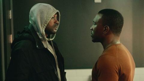 Kane "Kano" Robinson, who plays Sully, and Ashley Walters, who stars as Dushane in Top Boy. Sully and Dushane, two black men in their 20s, are pictured opposite and looking straight at each other with Sully on the left and Dushane on the right. Sully wears a grey hoodie with the hood pulled over his head beneath a black waterproof jacket. Dushane has short black hair and a moustache and wears an orange sweater. They are pictured inside with a grey wall behind them.