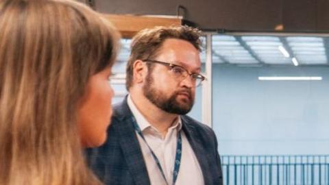 Matt Storey listens as someone speaks to him, we can also see the back of a young woman's head