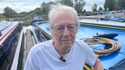 A man with grey wispy hair and a white t-shirt on, he has thin black framed  glasses and is leaning on the side of a narrow boat on the water, with other narrow boats around.
