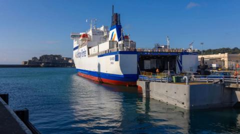 The Islander, a white ferry with red and blue trim, pulls up to a roll-on roll-off ramp in a harbour with a castle and tree-lined hills in the background on a sunny day. The back door of the ferry is open allowing vehicles to enter or leave.