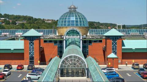 An orange fronted building with glass detail including a large glass dome. There is a sign in white writing which says 'Meadowhall' and surrounding the building is a car park with parked cars. 