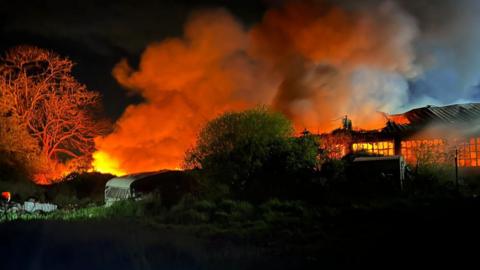 Black sky is permeated with grey smoke and flashes of orange from a fire. There is a tree with a caravan next to it in the middle of the photo with an industrial unit to the right. The unit is glowing orange through the windows.