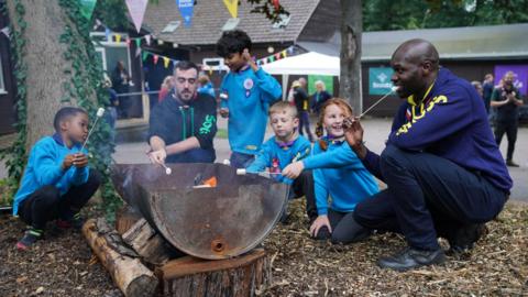 Chief Scout Dwayne Fields joins a scout leader with a moustache and four young people - three boys and a girl - in blue Beavers uniforms to toast marshmallows at a fire.