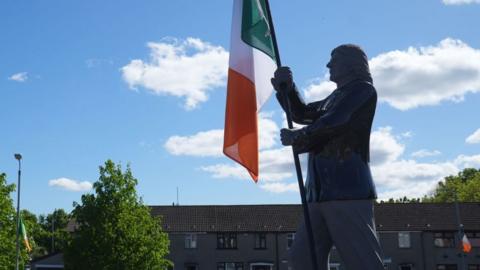 A metal statue of a long haired Bobby Sands, holding a green white and orange Irish flag, there is a row of brown, residential terraced houses in the bakcground and some large green trees