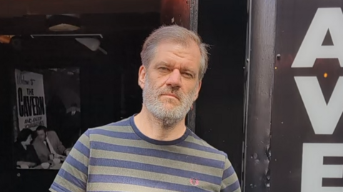A wide shot of David Dutton standing outside the Cavern Club. He has grey hair and is wearing a stripy t-shirt.