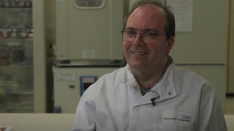 Toni Woodward wearing his working overalls wearing glasses and smiling at the camera. He is sitting in a lab.