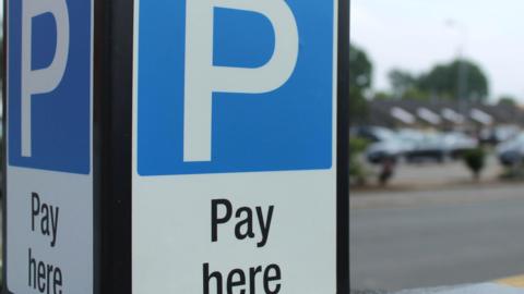 A blue and white parking sign with black lettering underneath saying"Pay here". Parked cars are out of focus in the background.