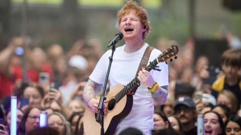 Ed Sheeran appears playing guitar, singing into a microphone, with people cheering behind him