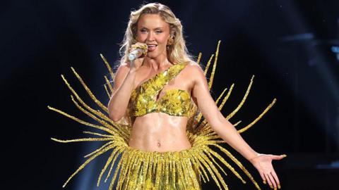 Zara Larsson performing onstage during the Grammy Awards in February. She is a white woman with blonde hair and is holding a shiny silver microphone in her right hand and signing into it with her mouth open. Her left arm is down with her palm outstretched. She is wearing a shiny yellow skirt which has lots of strands coming off it and going out from her side and back, as if inspired by a sun. She also has a cropped shiny yellow bra-style top on which has one strap across her shoulder.