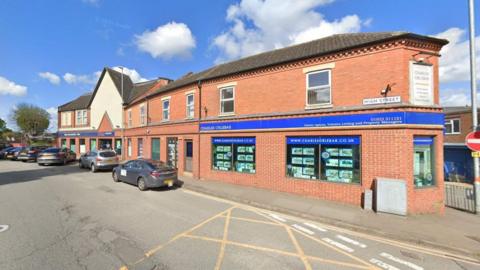 A brick building by the side of a road with several cars parked next to it. The blue branding of an estate agent is on the ground floor.