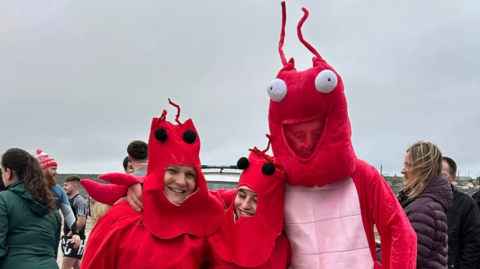 Three people wearing lobster fancy dress costumes standing on the beach with people walking around in the background.