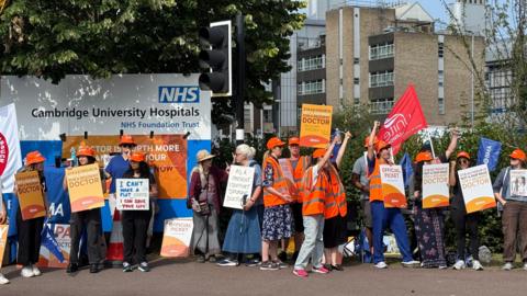 A group of protesters stand with placards and some wearing high-visibility jackets, in front of a sign that reads "NHS: Cambridge University Hospitals". They are holding placards supporting the doctors' strike and some have their hands in the air. 