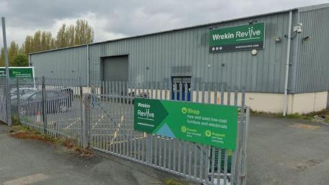 A single-storey corrugated metal warehouse building with its loading bay door shut and blue double doors, behind metal railings. Two cars are parked outside. A green sign on the railings points people towards Wrekin Reviive and a shop selling new and used low-cost furniture and electricals. A green Wrekin Reviive sign is also on the building.
