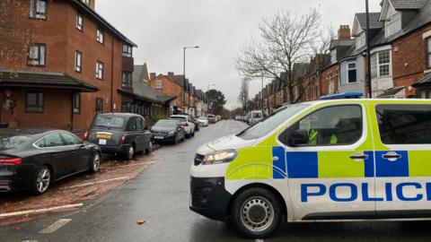 A police van is parked at the end of a road of terraced houses with cars parked either side