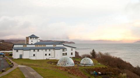 The college is a complex of white-walled buildings with grey roofs. The tallest is a turret like building. Next to the buildings are two small greenhouse-like glass domes. The site is on the coast and overlooks the sea.