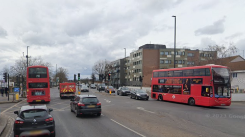 A StreetView image showing the four-way junction of Southbury Road and Baird Road in Enfield with various vehicles driving or waiting to turn, including red double-decker buses