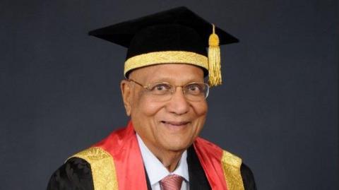 A man in a gradation cap and gown smiles at the camera. His gown has a red and gold hood lining and his cap is also lined with gold fabric.