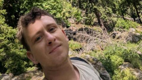 A man with short brown hair which is slightly curly on top taking a selfie surrounded by rocks, trees and general greenery. He is wearing a beige top and a black backpack.