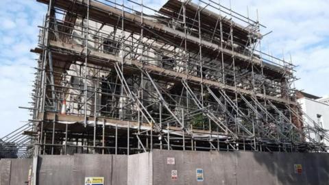 A grey building covered in a large quantity of scaffolding and surrounded by grey hoardings