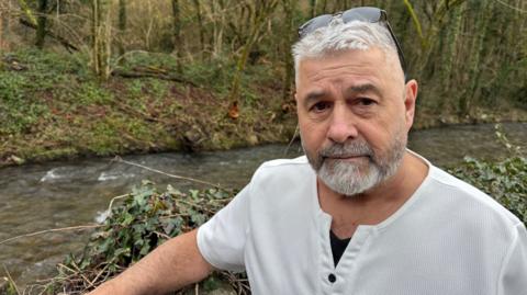 Man with grey hair, glasses resting on his head, standing by a brook and staring at the camera. He is not happy
