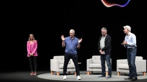 Apple CEO Tim Cook on a dark stage with a neon Apple logo behind him. He is wearing a blue polo sweater, black jeans and white sneakers and gesturing his hands, while three other people on stage look at him.
