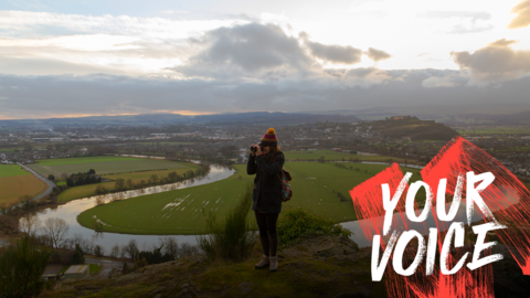A woman is standing on top of her grassy hill. Behind her is the large meander of a river and the Scottish countryside. The sun is trying to break through the clouds. 