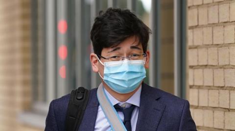 Jonathon Dean wearing a navy suit with a navy tie and a white shirt and glasses. He is walking outside of a court building with the strap of a black rucksack over one shoulder and a facemask on.