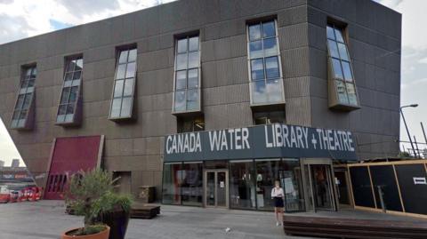 Inverted pyramid building with 'Canada Water Library and Theatre' sign on the right hand side. A woman stands in the foreground.